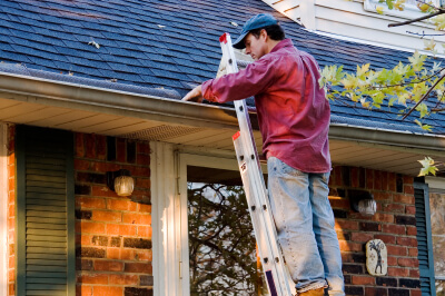 Man on ladder cleans leaves out of his gutter during seasonal home maintenance.